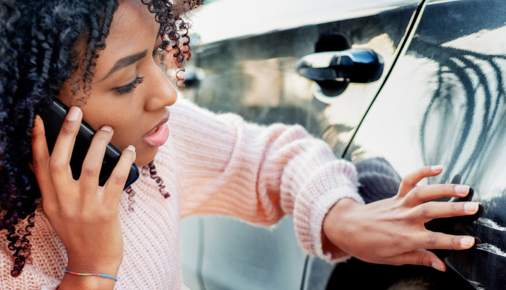 A woman holding her mobile phone to her ear while examining a scratch on a car’s paintwork.