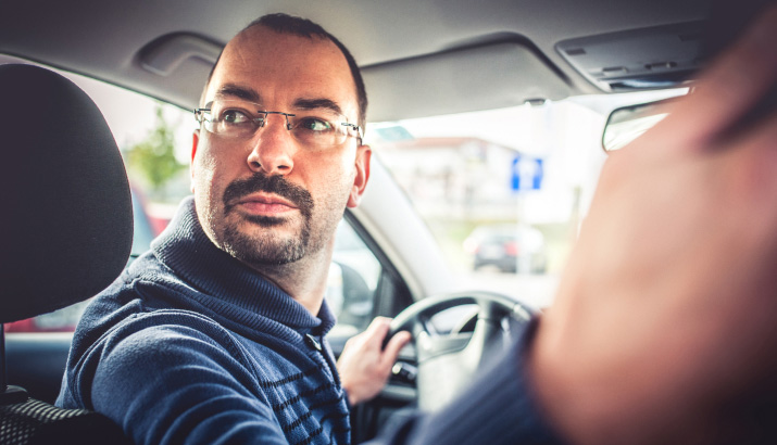 A man holding the steering wheel and looking behind as he reverses his car.