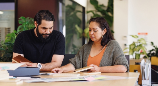 A man and woman examine work material at a table with plants in the background