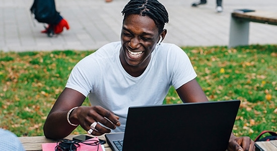 A young man with a bright smile works on a laptop outdoors