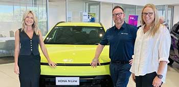 Karlie Male stands on the lefthand side of a yellow Hyundai Kona, while Aleesha Van Vuuren, Allianz Account Manager, and James Liddiard, Wangara Hyundai Business Manager stand on the righthand side of the vehicle inside a Hyundai motor dealership.