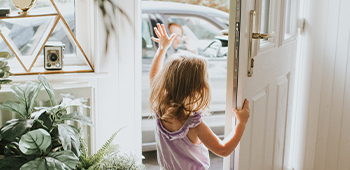 Young girl waves through the front door at a driver who waves back from inside their vehicle in the driveway.