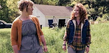 Two women in a rural setting walking across green grass and smiling at each other
