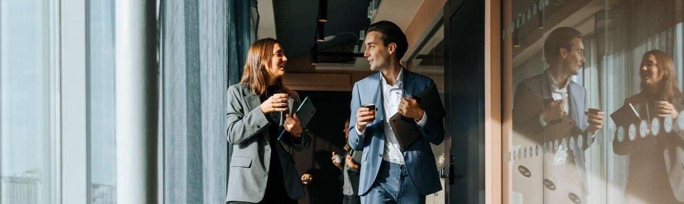 A woman and man hold coffees as they have a conversation in an office corridor