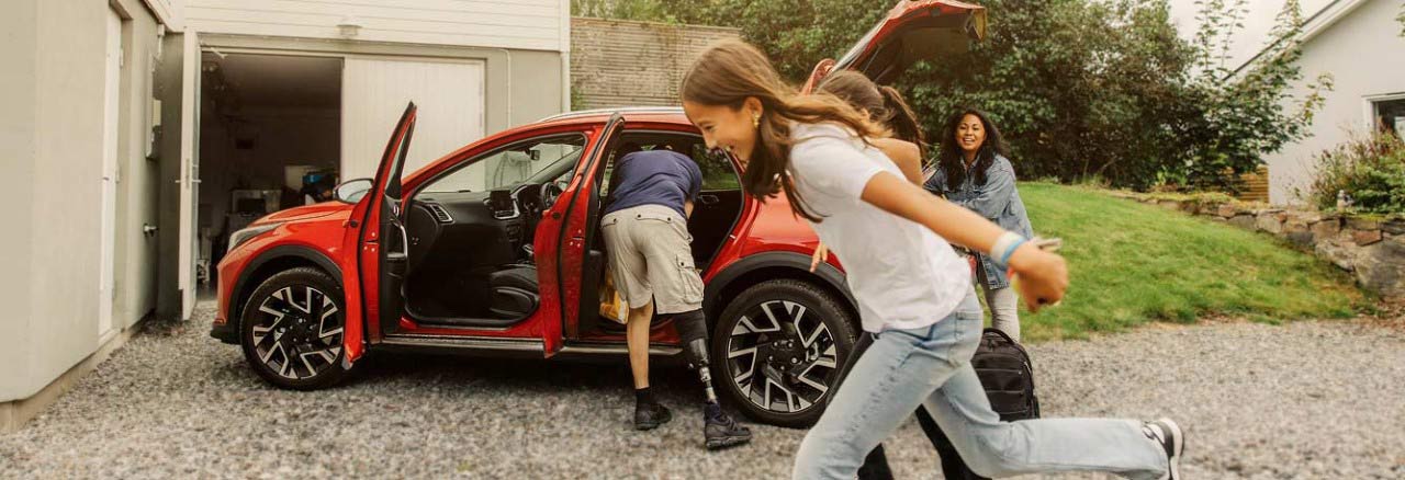 A family loading a car in a driveway, with children running and a woman smiling.