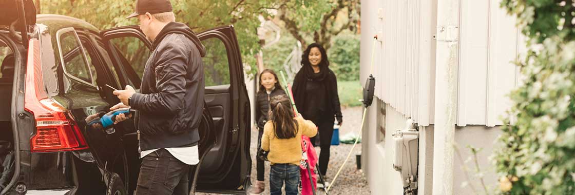 Father charging an electric vehicle while wife and children load items into the boot ahead of a trip