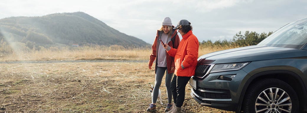 A couple standing next to their broken-down car, using a mobile phone to call for help.