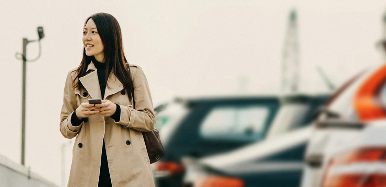 A woman in a beige trench coat holds a smartphone with parked cars in the background