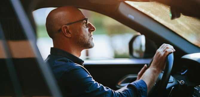 Person driving a car, viewed from the side, with hands on the steering wheel.