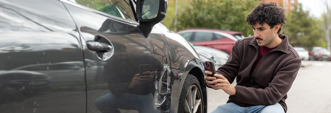 A man crouches next to a car with a dent, taking a photo with his smartphone in a parking lot.