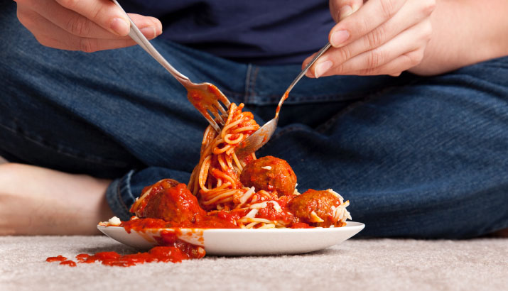 Someone sitting on the floor to eat a plate of spaghetti, with sauce spilled on the carpet.