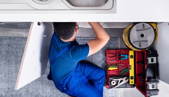 A plumber working below a sink next to an open box of tools.