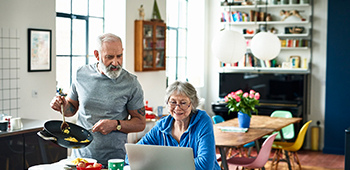 A man holds a pan of eggs while looking at his wife’s laptop