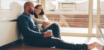 Heterosexual middle-aged couple seated leaning against a wall in casual conversation holding coffee mugs