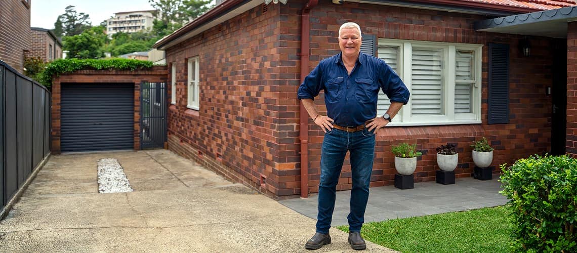 Scotty Cam stands in front of a well-maintained house with his hands on his hips. 