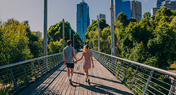 A couple walking hand in hand across a bridge with trees on either side and a cityscape ahead.