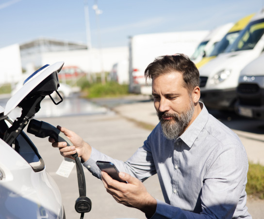 Middle aged man charging his electric vehicle and using his phone, with vans and a warehouse in the background.