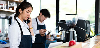 A female coffee shop barista is taking an order on smart phone and stands behind the cashier counter while a male barista makes coffee in the background.