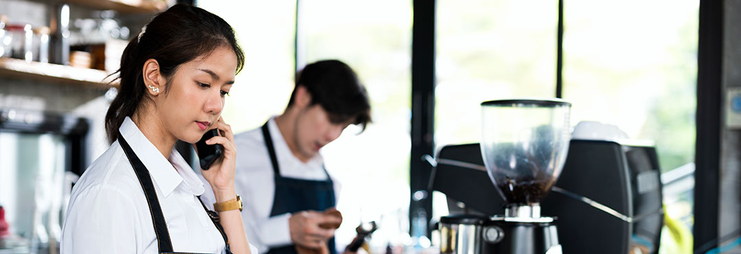 A Female coffee shop barista is taking an order on smart phone and stands behind the cashier counter while a male barista makes coffee in the background.